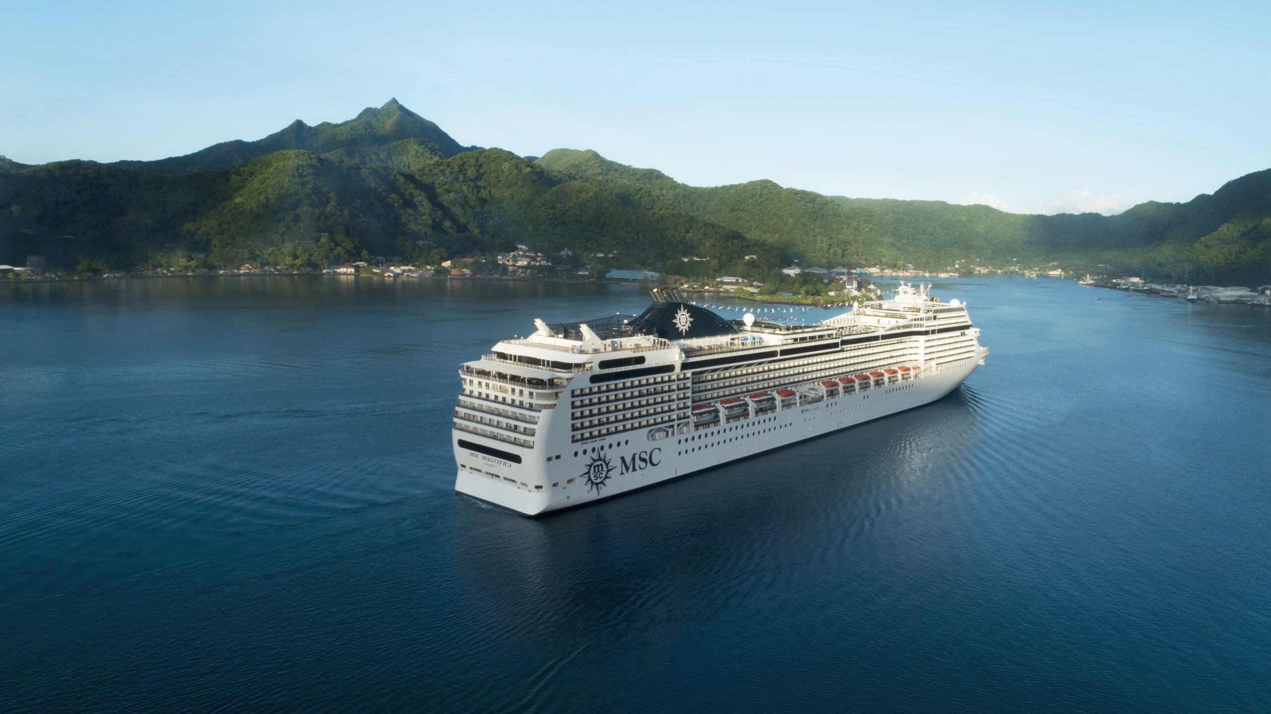 The MSC World America, a large, modern white cruise ship with the MSC logo visible on its hull, cruising on calm turquoise water surrounded by lush green mountainous islands under a clear blue sky.