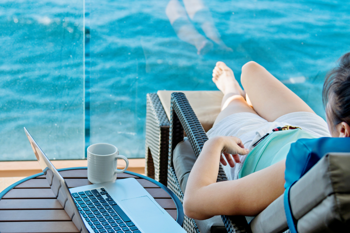 A woman on a cruise ship balcony working on a laptop, enjoying a cup of coffee, and practicing cruise wellness.